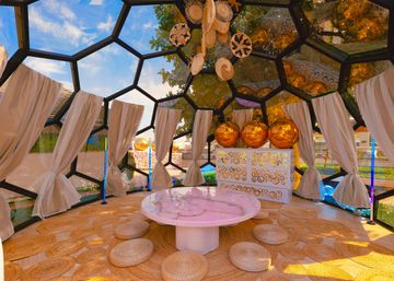 Boho-style geodesic glass dome lounge in a garden with tied linen curtains, woven round floor cushions and mats, a low pink marble table, hanging wicker decor and large reflective golden orb lights.
