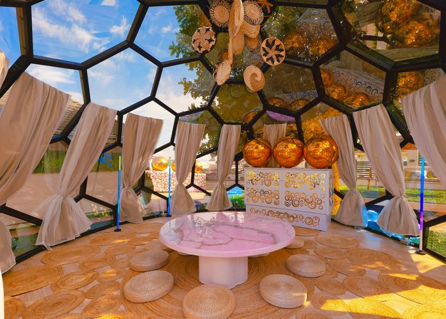 Boho-style geodesic glass dome lounge in a garden with tied linen curtains, woven round floor cushions and mats, a low pink marble table, hanging wicker decor and large reflective golden orb lights.