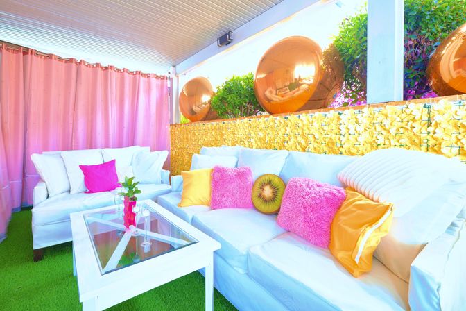 Bright outdoor patio lounge with white sofas on green artificial turf, pink curtains, colorful throw pillows (pink, yellow, kiwi-shaped), glass coffee table, and reflective gold decorative spheres along a floral-textured wall.