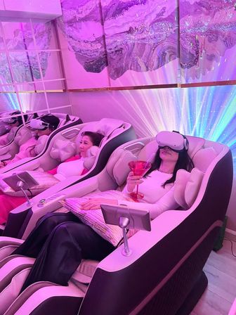 Women reclining in VR headsets inside luxury massage chairs in a pink-lit wellness lounge, one holding a cocktail amid colorful LED wall art and ambient light effects.