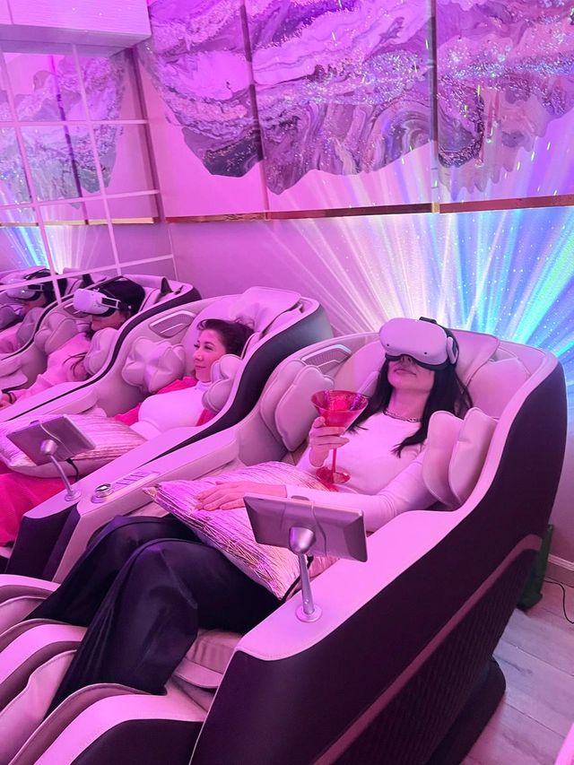 Women reclining in VR headsets inside luxury massage chairs in a pink-lit wellness lounge, one holding a cocktail amid colorful LED wall art and ambient light effects.