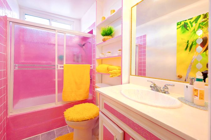 Cheerful pink residential bathroom with frosted pink shower doors, white vanity sink, yellow towel and fuzzy yellow toilet seat cover, open shelves with stacked yellow towels and a small green plant.