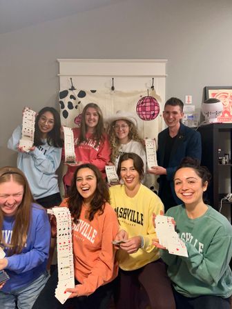 Smiling group of friends indoors holding long cascading decks of playing cards, wearing colorful casual sweatshirts (some reading “Nashville”) and posing in front of a playful backdrop with a pink disco ball illustration.