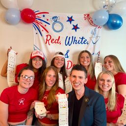 Group of smiling people in red shirts holding long fanned playing-card decks in front of a red, white and blue celebratory banner and balloons