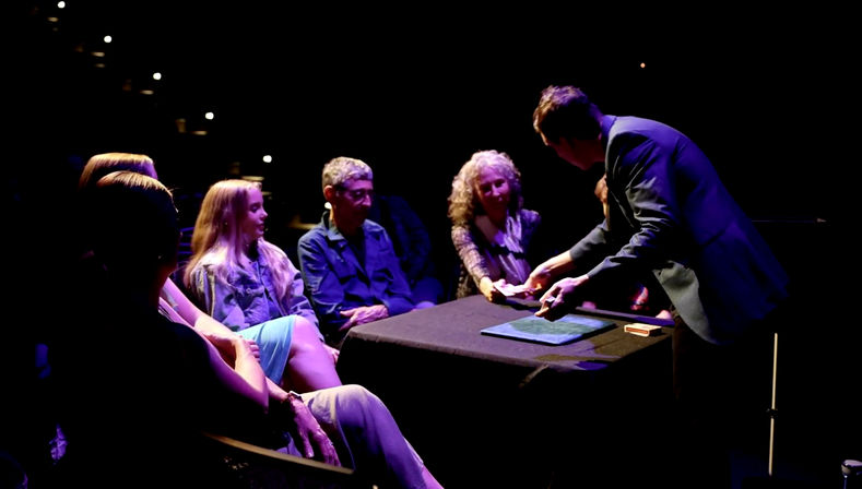 Magician performing a close-up card trick at a small theater table, handing cards to an attentive seated audience under purple-blue stage lighting.