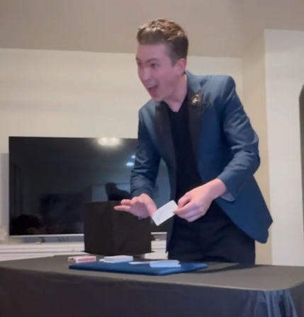 Entertaining magician in a blue blazer doing a lively card trick over a black-covered table with decks of playing cards in a living room setting.