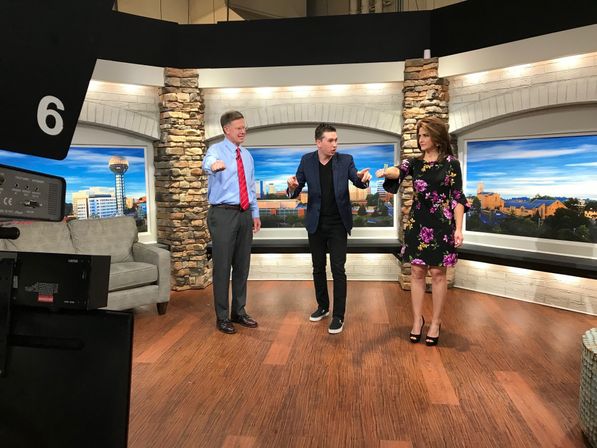 Three on-air hosts on a Knoxville morning show set with stone pillars and a city skyline backdrop (Sunsphere visible); a man in a blue shirt and red tie, a man in a dark blazer, and a woman in a black floral dress gesture toward the camera, studio camera and couch in foreground.