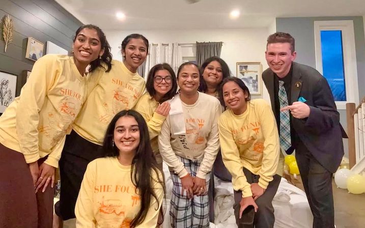 Group photo of eight friends in a cozy living room — most wearing matching yellow sweatshirts, smiling and posing during a casual indoor celebration with balloons.