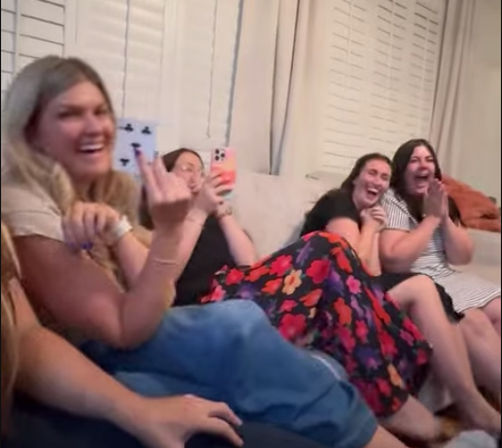 Group of friends laughing on a living-room couch during a cozy get-together, one holding a smartphone, colorful floral skirt and white shutters in the background.