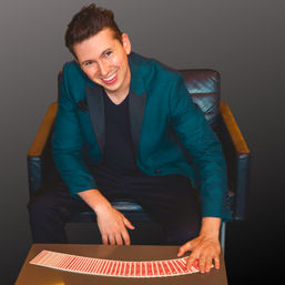 Cheerful magician in a teal blazer seated in a leather chair fans a deck of red-backed playing cards across a table in a studio portrait