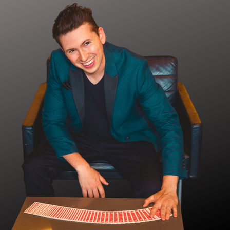Cheerful magician in a teal blazer seated in a leather chair fans a deck of red-backed playing cards across a table in a studio portrait