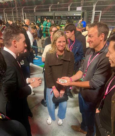 Magician in a suit performing a card trick for a small group of smiling spectators with pink VIP lanyards in a crowded nighttime racetrack pit lane, barriers and crew in green uniforms visible in the background.