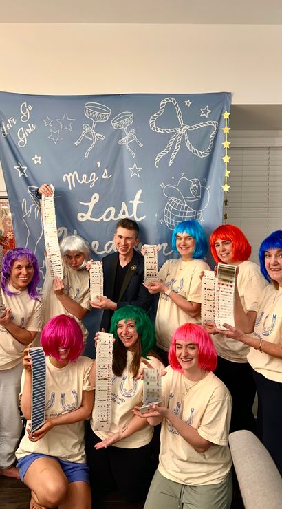 Bachelorette party group photo in a living room: eight people in matching t-shirts and bright colorful wigs smiling and holding long cascading decks of playing cards in front of a blue celebratory banner.