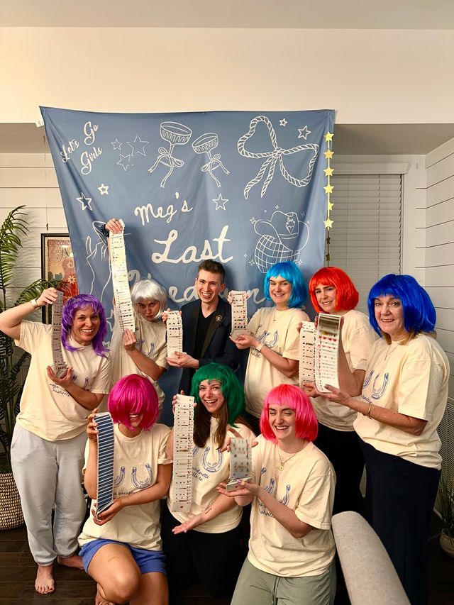 Bachelorette party group photo in a living room: eight people in matching t-shirts and bright colorful wigs smiling and holding long cascading decks of playing cards in front of a blue celebratory banner.