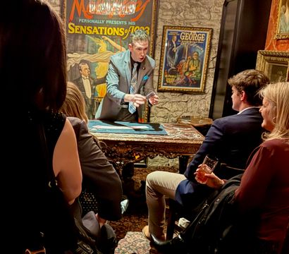Close-up magician with dramatic expression performing a card trick for a seated audience in an intimate vintage-style room with an ornate table, framed retro posters and stone wall backdrop — live close-up magic show scene.