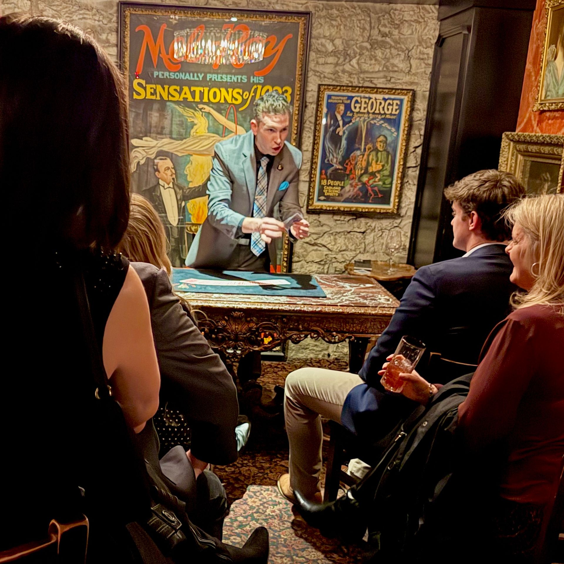 Close-up magician with dramatic expression performing a card trick for a seated audience in an intimate vintage-style room with an ornate table, framed retro posters and stone wall backdrop — live close-up magic show scene.