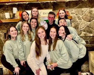 Group of friends in matching mint sweatshirts smiling and posing in a cozy cabin living room with a stone fireplace during an indoor celebration