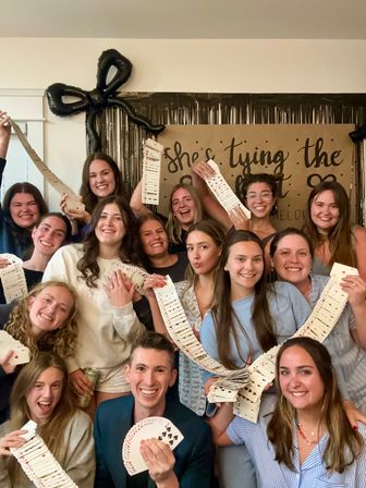 Smiling group photo at an indoor party, friends holding long cascading decks of playing cards in front of a kraft paper backdrop, black bow balloon and metallic fringe decorations — playful bachelorette-style celebration.