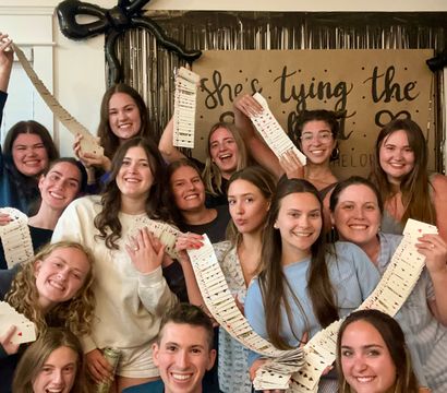 Smiling group photo at an indoor party, friends holding long cascading decks of playing cards in front of a kraft paper backdrop, black bow balloon and metallic fringe decorations — playful bachelorette-style celebration.