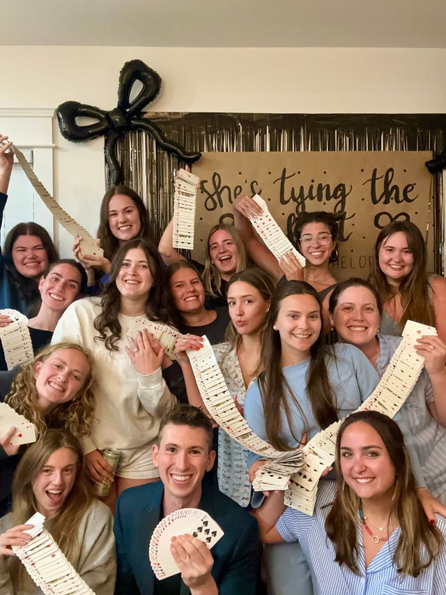 Smiling group photo at an indoor party, friends holding long cascading decks of playing cards in front of a kraft paper backdrop, black bow balloon and metallic fringe decorations — playful bachelorette-style celebration.