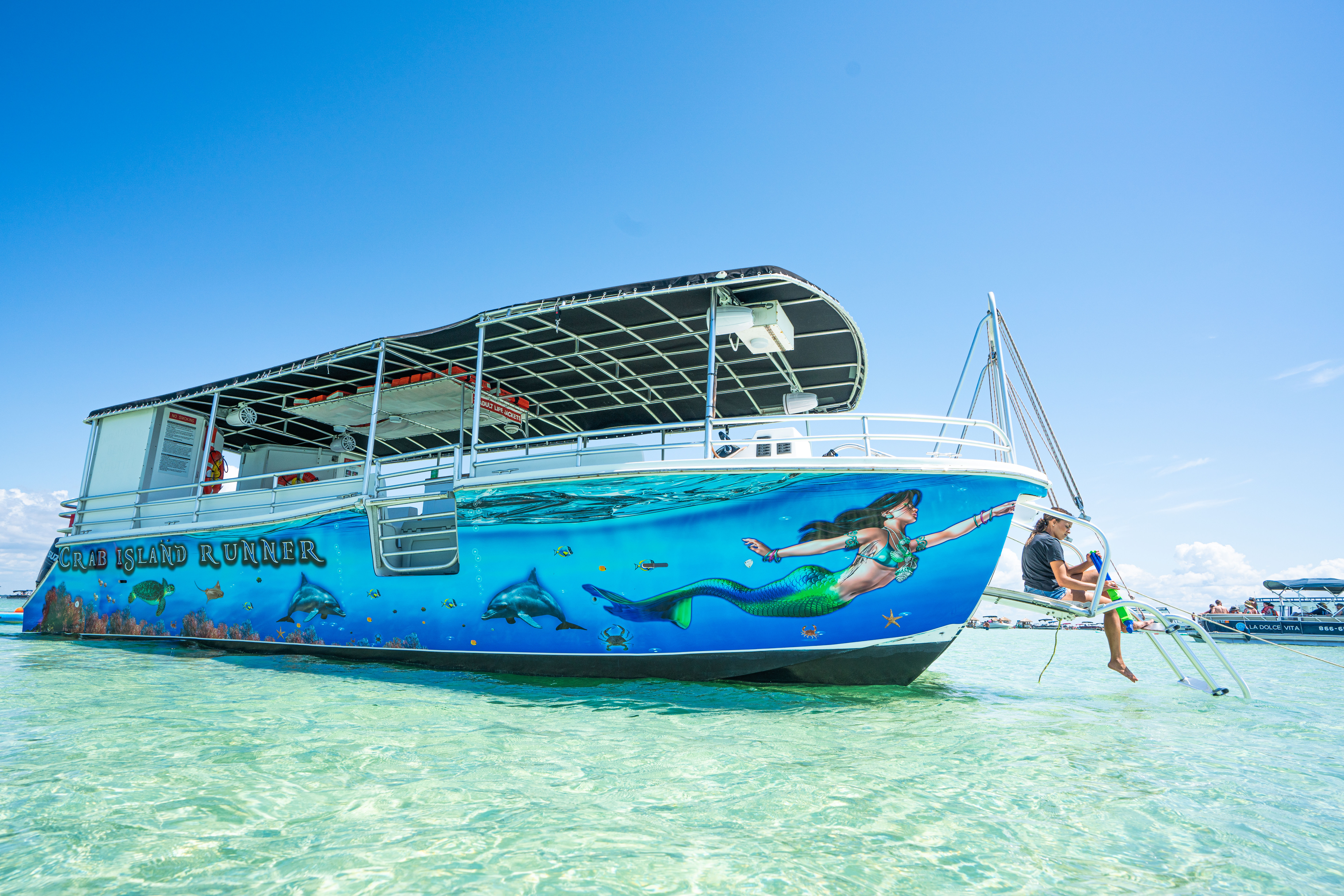 Colorful pontoon boat with a mermaid and dolphin mural anchored in clear turquoise shallow water at a tropical sandbar, a person sitting on the swim ladder under a bright blue sky
