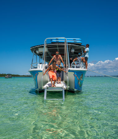 People stepping off a colorful tour boat into crystal-clear turquoise shallow water at a sunny sandbar, with kayaks stacked on the roof and fish artwork on the hull.