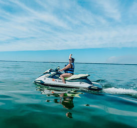 Person in a blue life jacket riding a white-and-green jet ski on calm turquoise coastal waters, raising one arm beneath a bright blue sky with wispy clouds and a clear reflection on the water.