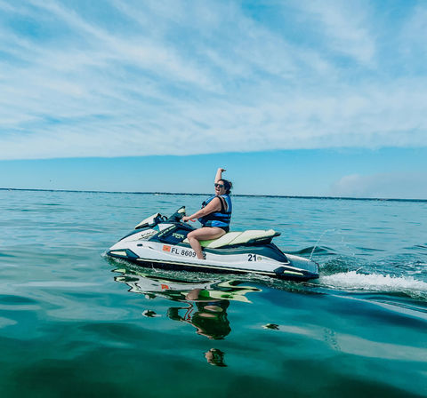 Person in a blue life jacket riding a white-and-green jet ski on calm turquoise coastal waters, raising one arm beneath a bright blue sky with wispy clouds and a clear reflection on the water.