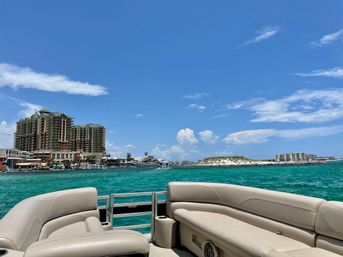 View from a pontoon boat over turquoise coastal waters toward a beachfront resort skyline, sandy island and marina under a bright blue sky — sunny vacation vibes.
