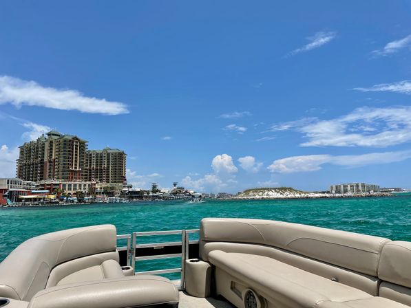View from a pontoon boat over turquoise coastal waters toward a beachfront resort skyline, sandy island and marina under a bright blue sky — sunny vacation vibes.