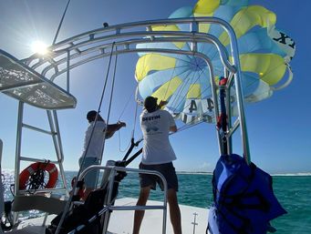 Two crew members on a parasailing boat launching a yellow-and-blue parachute over turquoise coastal waters beneath a bright sunny sky.