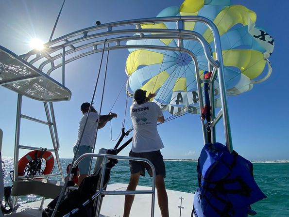 Two crew members on a parasailing boat launching a yellow-and-blue parachute over turquoise coastal waters beneath a bright sunny sky.