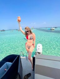 Woman in red-and-leopard bikini raising a drink on a boat ladder over crystal-clear turquoise water with anchored boats on a sunny tropical day