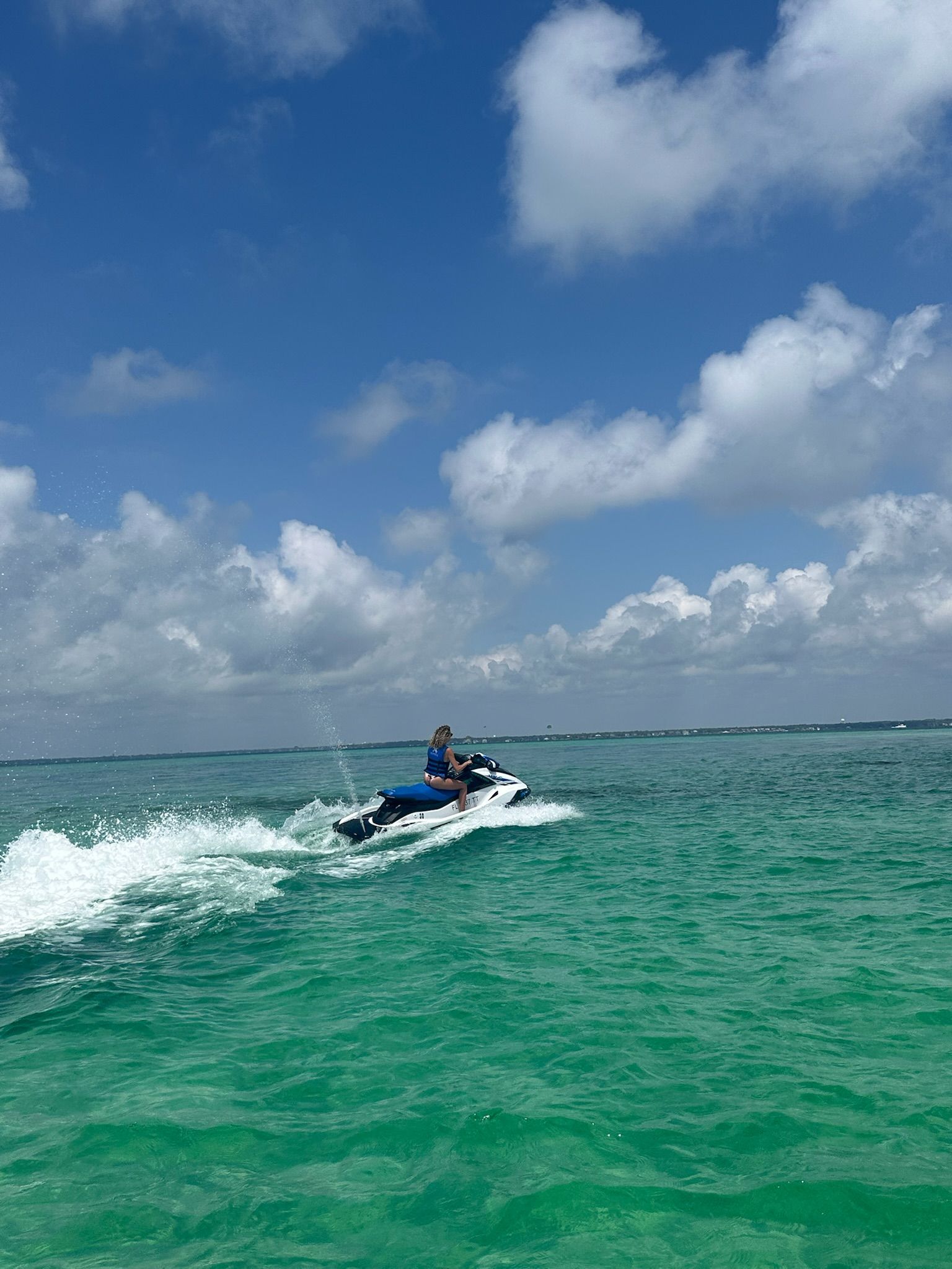 Person wearing a blue life jacket riding a white jet ski across turquoise coastal waters, kicking up a foamy wake under a bright blue sky dotted with puffy clouds.