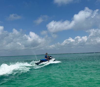 Person wearing a blue life jacket riding a white jet ski across turquoise coastal waters, kicking up a foamy wake under a bright blue sky dotted with puffy clouds.