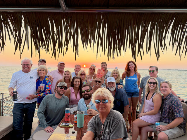 Large group of adults smiling and holding drinks under a thatched tiki canopy on a boat at a colorful ocean sunset — tropical sunset cruise/boat party scene.