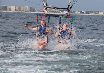 Two people in blue life jackets laughing as their tandem parasail splashes into the ocean with beachfront condos on the horizon