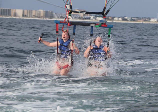 Two people in blue life jackets laughing as their tandem parasail splashes into the ocean with beachfront condos on the horizon