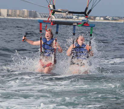 Two people in blue life jackets laughing as their tandem parasail splashes into the ocean with beachfront condos on the horizon