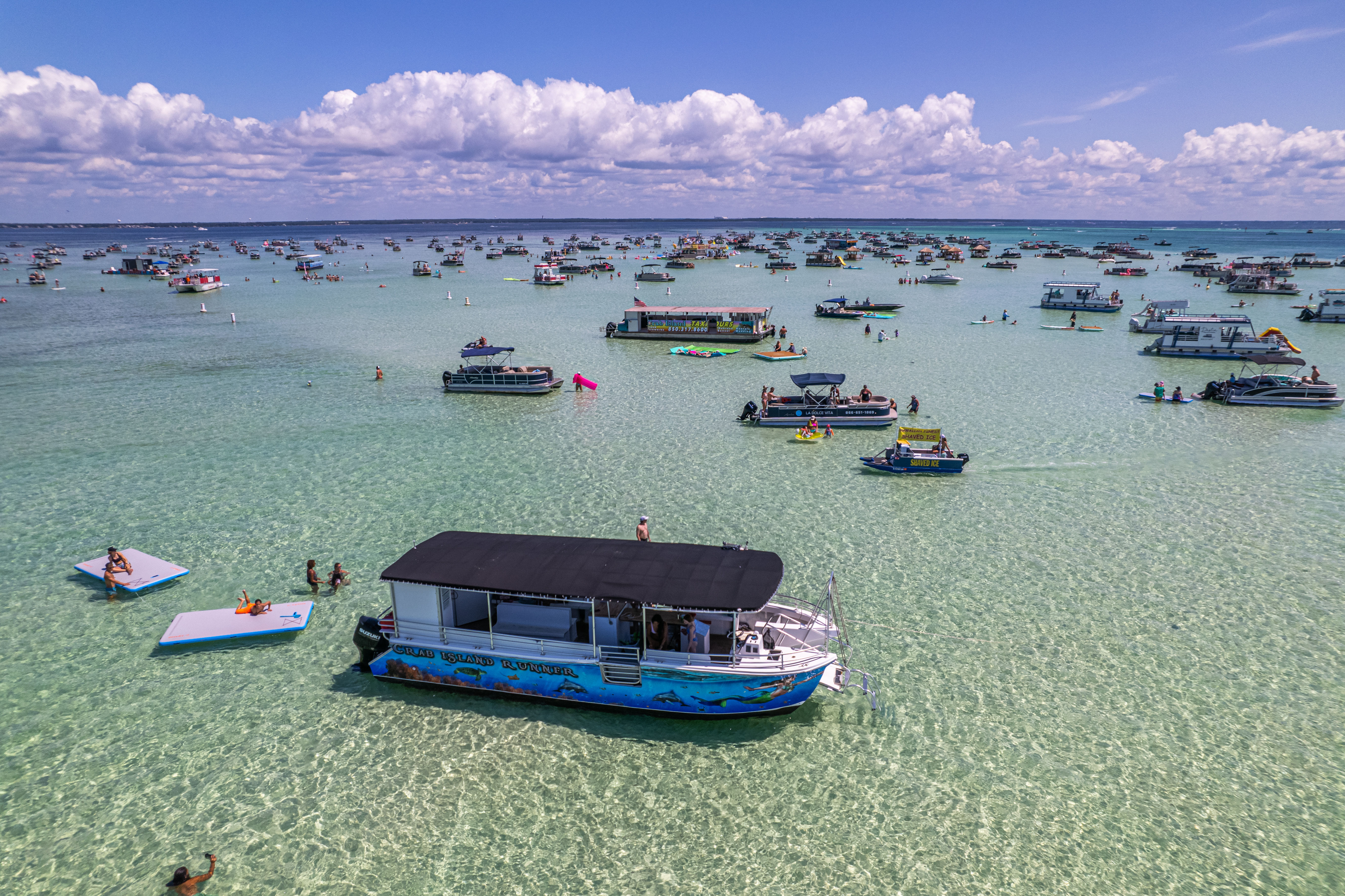 Drone view of a crowded Florida sandbar with dozens of anchored pontoon boats, people swimming and lounging on floating platforms in crystal-clear shallow turquoise water under a blue sky with puffy clouds.