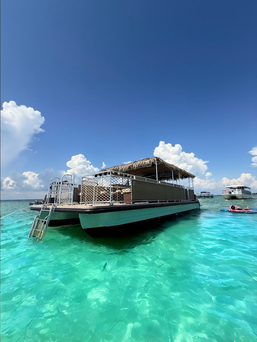 Thatched-roof pontoon boat with ladder floating on clear turquoise tropical water under a bright blue sky with puffy clouds.