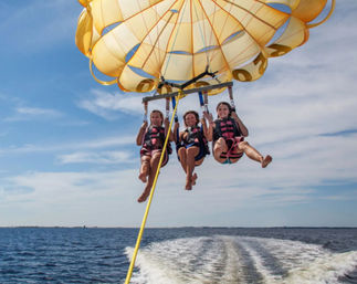 Three people parasailing under a bright yellow parachute over blue ocean waters, hovering above a boat wake beneath a sunny sky.