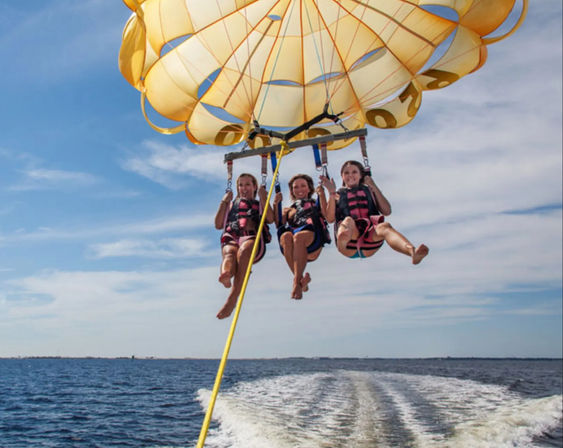 Three people parasailing under a bright yellow parachute over blue ocean waters, hovering above a boat wake beneath a sunny sky.