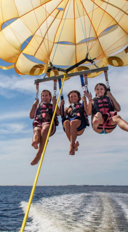 Three people parasailing under a bright yellow parachute over blue ocean waters, hovering above a boat wake beneath a sunny sky.
