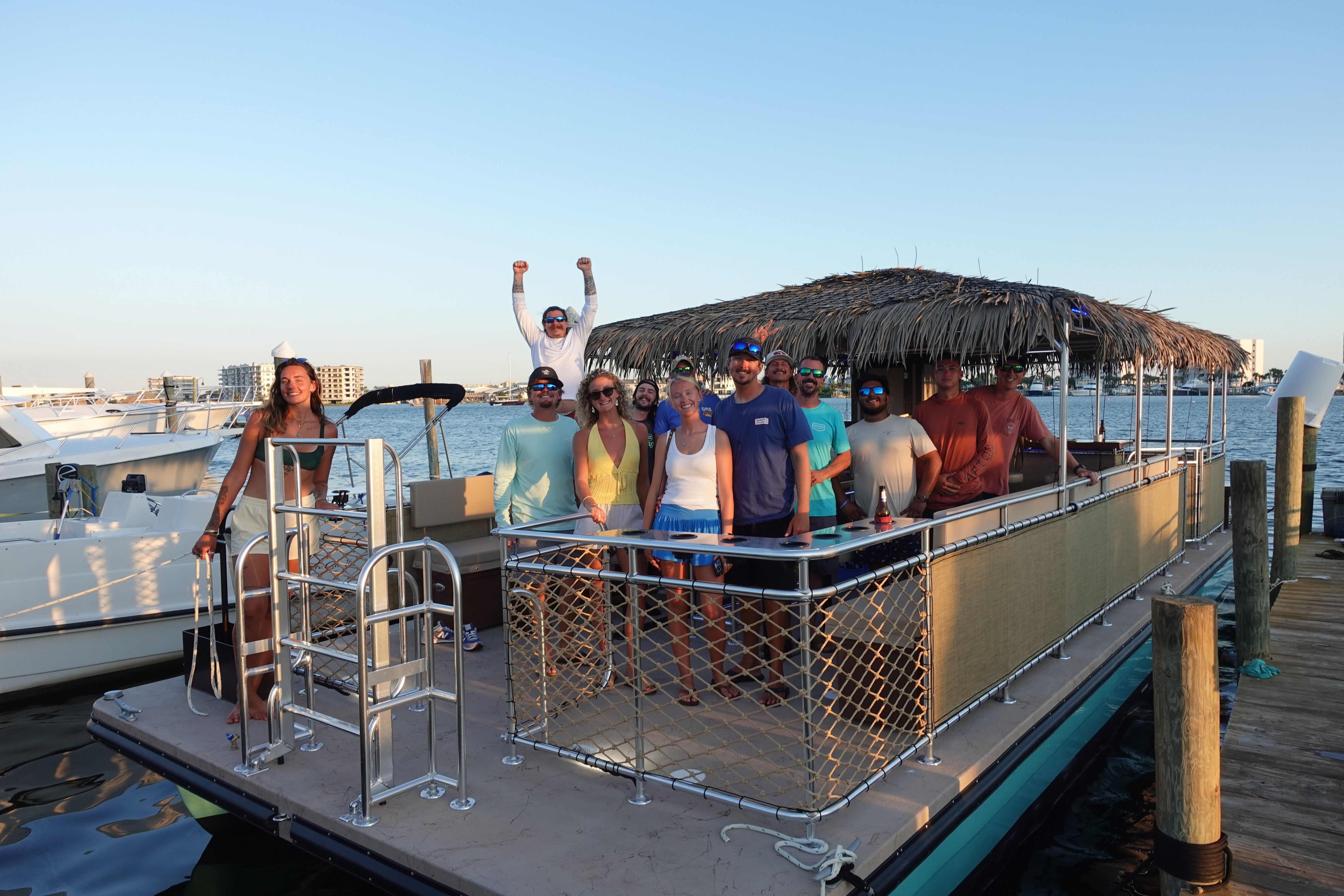Group of people on a tiki-style pontoon boat docked at a coastal marina, smiling and enjoying a sunny sunset cruise