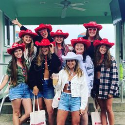 Smiling group of friends in red cowboy hats and denim shorts at a seaside pavilion, bride-to-be in a white hat holding a drink — beach bachelorette celebration