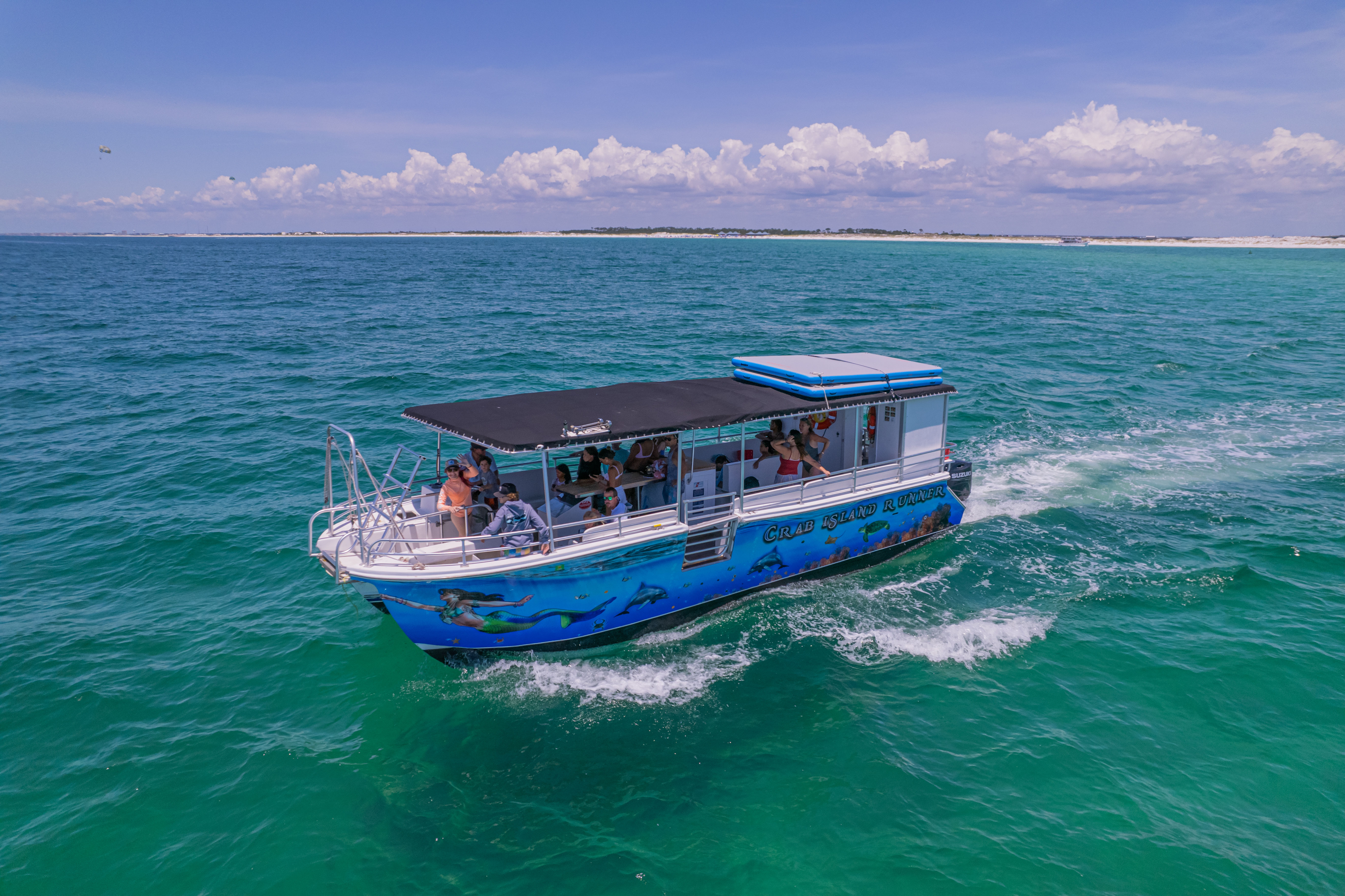 A vibrant pontoon tour boat with passengers cruising across emerald-turquoise ocean, leaving a white wake with a sandy shoreline and fluffy clouds on a bright blue sky — aerial coastal view.