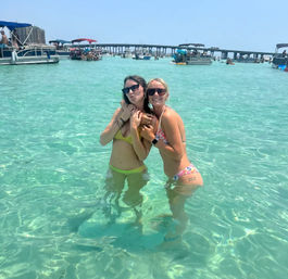 Two smiling women in colorful bikinis and sunglasses standing waist-deep in clear turquoise water near a busy sandbar with pontoon boats and a low bridge on a sunny day.