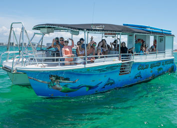 Passengers waving from a colorful mermaid- and dolphin-painted tour boat on clear turquoise coastal waters under a sunny sky