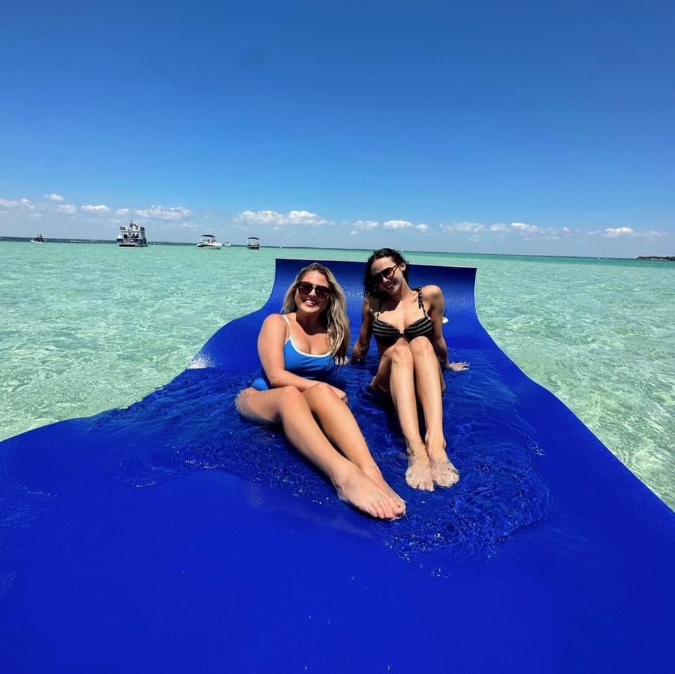 Two smiling women in swimsuits lounging on a bright blue floating mat in shallow turquoise water with anchored boats on the horizon and a clear sunny sky